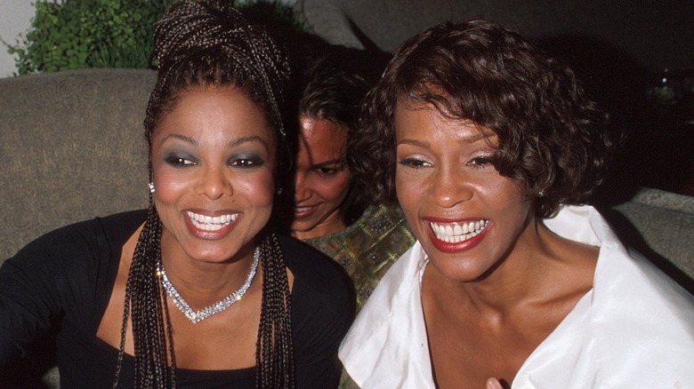 Whitney Houston sentada y sonriendo junto a Janet Jackson