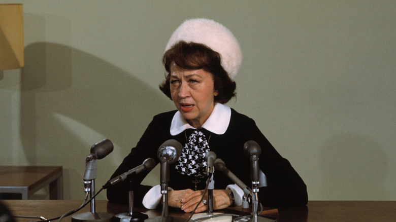 Jeane Dixon hablando en micrófonos, con sombrero peludo