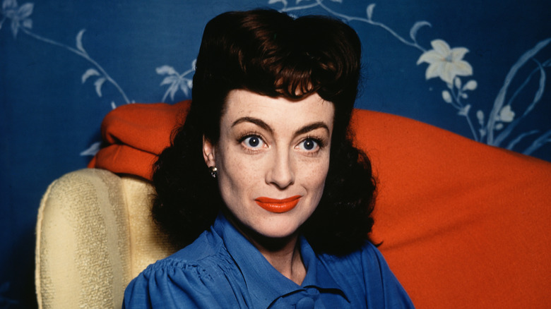 Joan Crawford en una foto en interiores, sentada en una silla con fondo de papel de flores azul