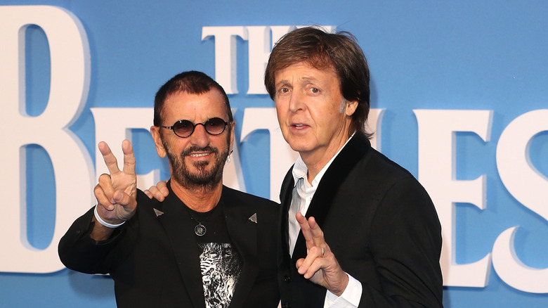 Ringo Starr haciendo la señal de paz junto a Paul McCartney frente al logo de los Beatles