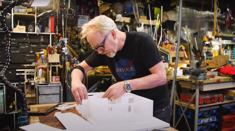 Adam Savage con barba y gafas en su taller haciendo una maqueta de su casa