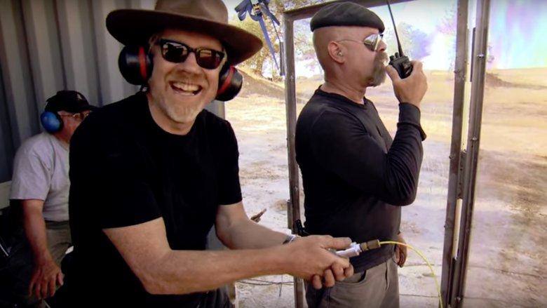 Adam Savage con barba y gafas de sol sonriendo junto a Jamie Hyneman en un campo de tiro