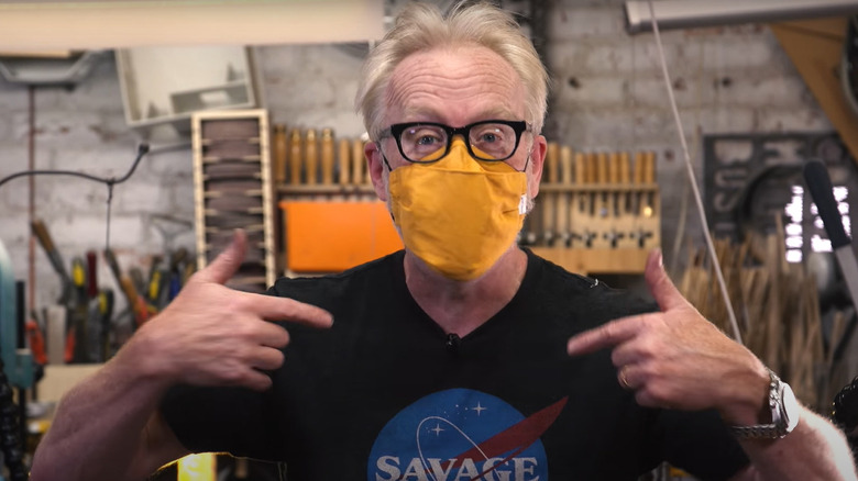 Adam Savage con gafas y mascarilla amarilla señalándose a sí mismo en taller