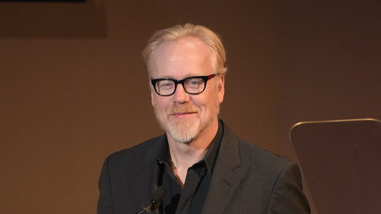 Adam Savage sonriendo con barba y gafas en traje oscuro